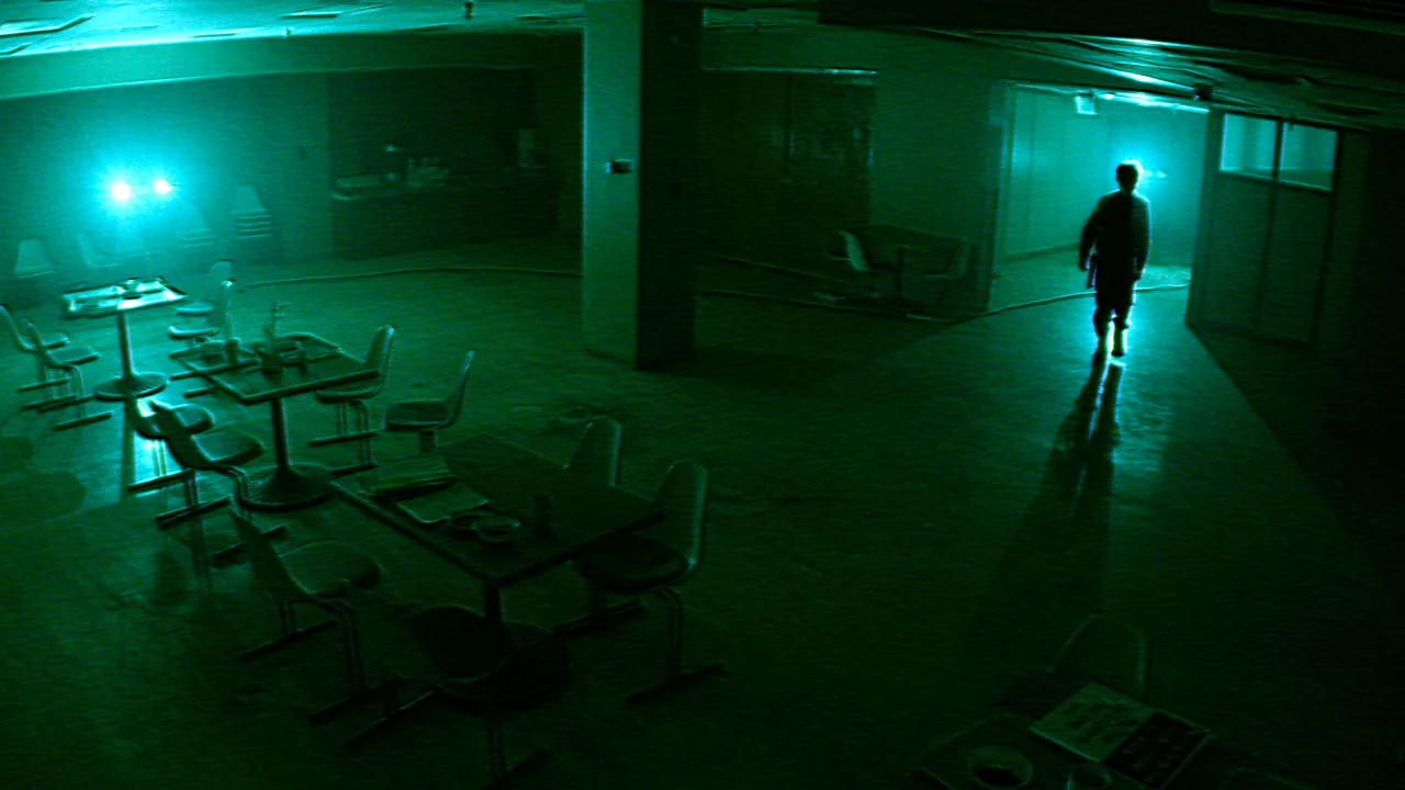 Imagen promocional de Infirmary: vista elevada de una sala oscura con mesas y una figura caminando por el hospital